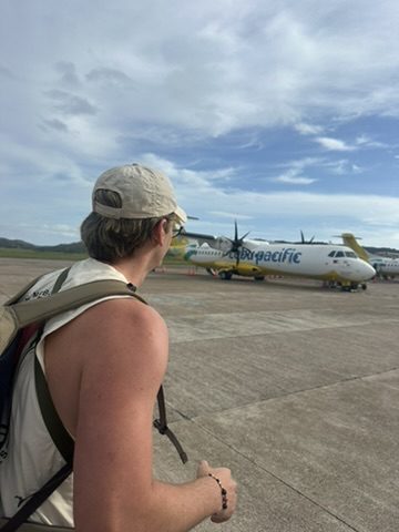 Ryker at the tarmac of a small Philippine airport looking at a Cebu Pacific propeller plane