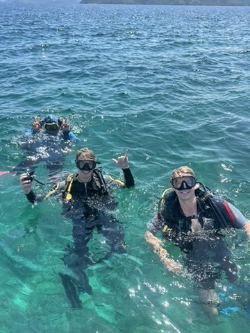 Three divers in crystal-clear shallow water giving shaka signs near the wreck site