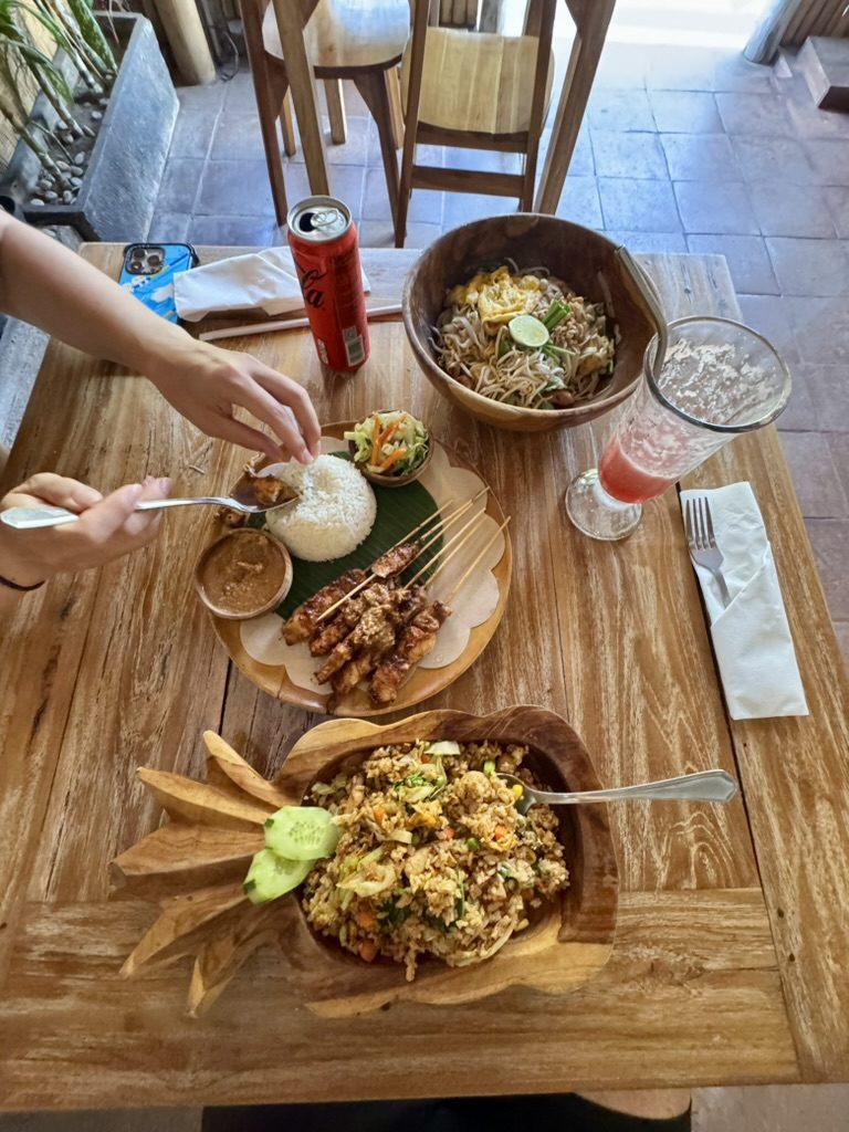 Overhead shot of a Balinese food feast — chicken sate with peanut sauce, nasi goreng, mie goreng, and fresh juice