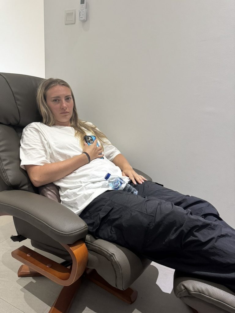 Meg slumped in a clinic recliner looking exhausted and pale, clutching a water bottle