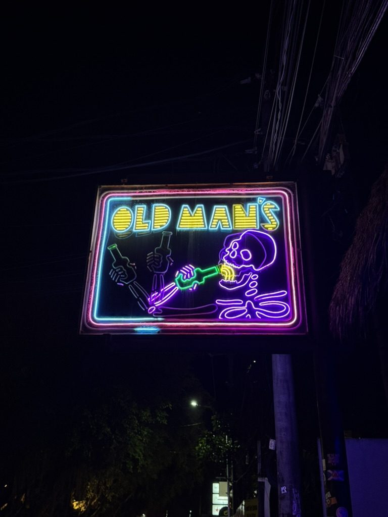 Old Man's bar neon sign at night — a skeleton drinking from a bottle in vivid neon colors