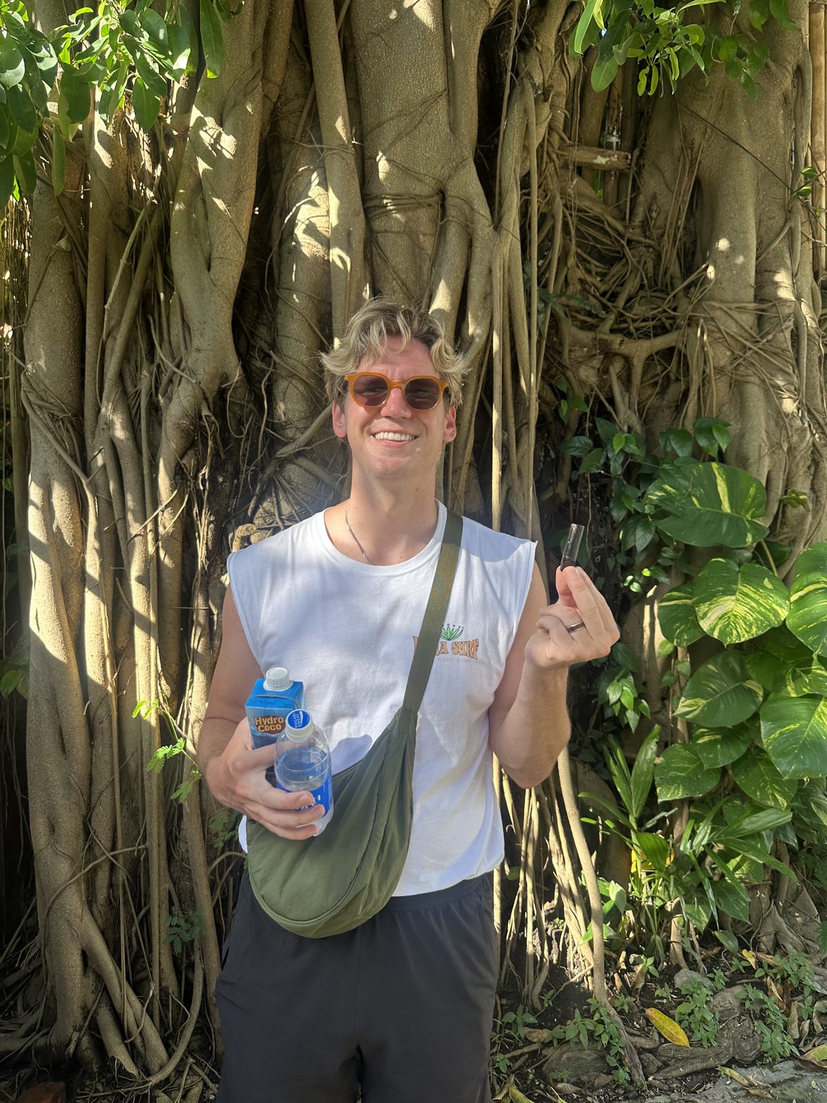 Ryker holding charcoal pills triumphantly in front of a massive Bali banyan tree, holding a Hydro Coco