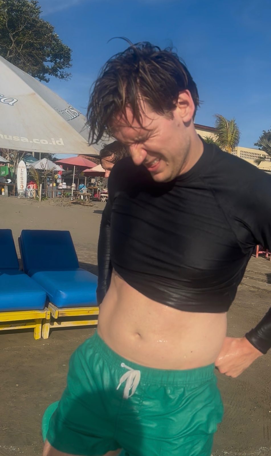 Ryker peeling off a soaking wet black rash guard on the beach after surfing in the dirty water