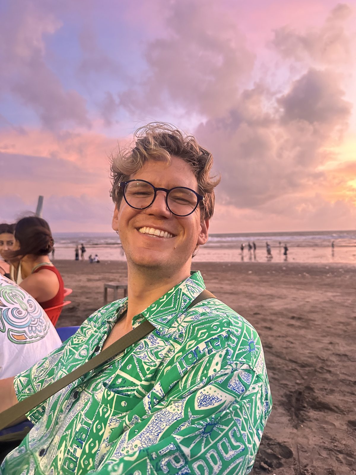 Ryker grinning ear to ear at the Bali sunset in his green patterned shirt, beach behind him
