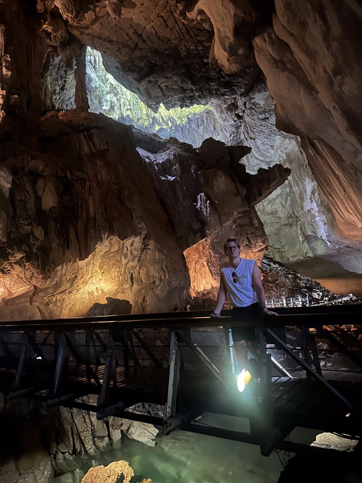Ryker on the illuminated cave boardwalk above a green glowing underground river, light shaft pouring in from above
