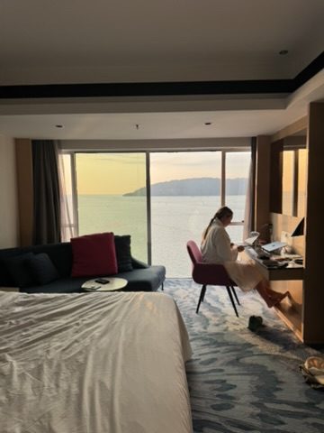 Meg sitting at the Le Méridien desk in a white robe, South China Sea sunset glowing through the floor-to-ceiling window