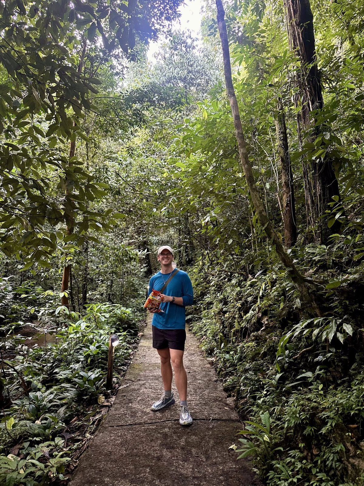 Ryker smiling on a jungle trail surrounded by towering Borneo rainforest, holding a snack bag