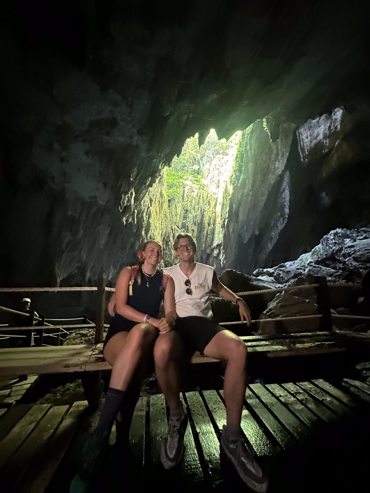Meg and Ryker sitting on a cave boardwalk with a dramatic shaft of light and green jungle visible through the rock opening above